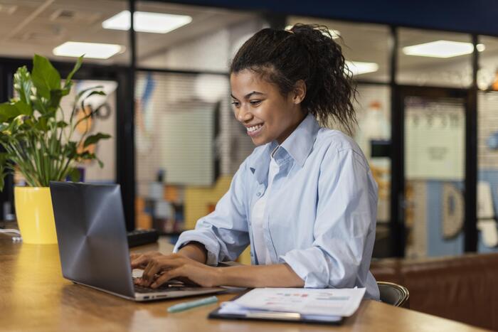 Mulher sorrindo enquanto trabalha no notebook dentro de um ambiente corporativo moderno, representando a locação de sala por hora para profissionais que precisam de espaços flexíveis e equipados. 