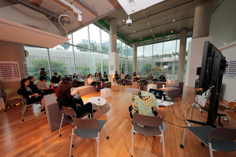 Workshop em andamento em sala ampla, espaçosa, com diversas cadeiras, sofás, televisão.